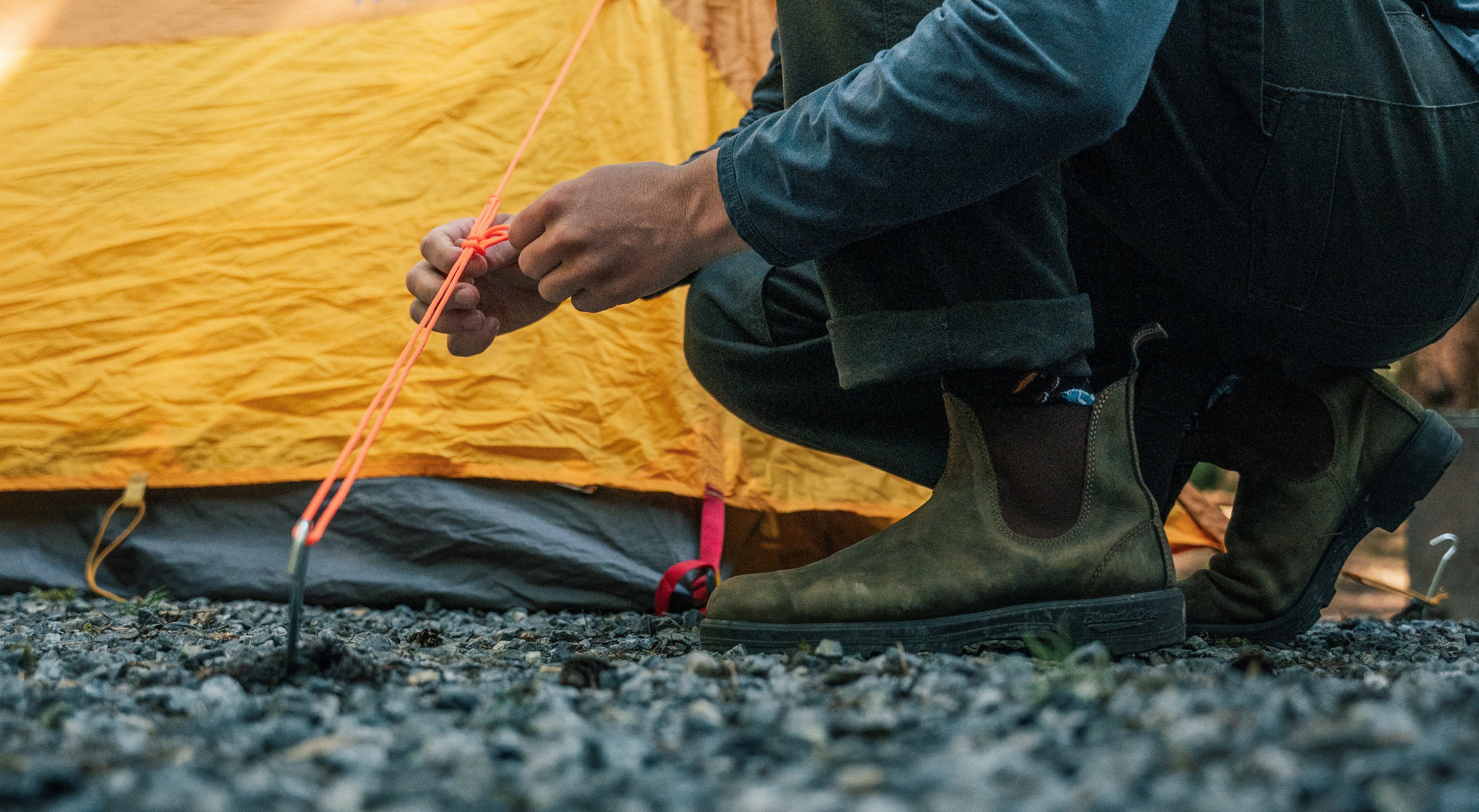 Close up of a man crouched in front of a tent tying a knot with orange rope